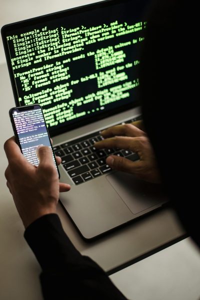 ciberseguridad-programacion-movil Persona trabajando en ciberseguridad con un portátil y un móvil, código verde en pantalla, manos tecleando.